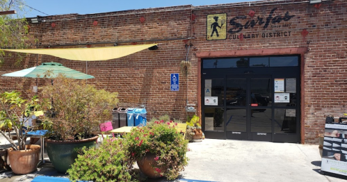 Entrance to a rustic brick building with outdoor seating, plants, and umbrellas, labeled "Culinary District."