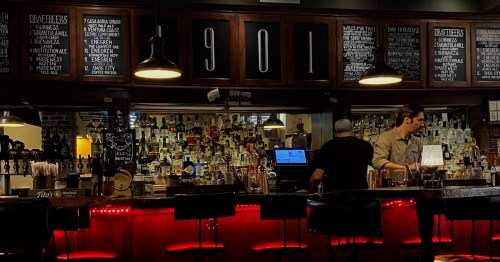 A dimly lit bar with a wooden counter, shelves of liquor, and a bartender preparing drinks.