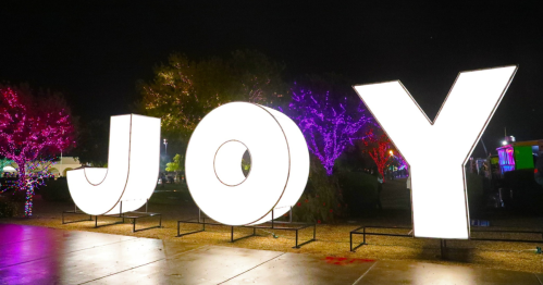 Large illuminated letters spelling "JOY" in a festive outdoor setting with colorful lights on trees.