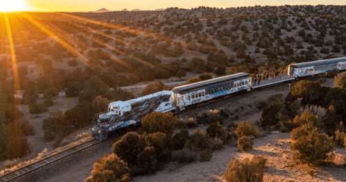 A colorful train travels along a winding track through a desert landscape at sunset, with rays of sunlight shining.