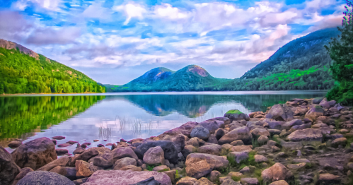 A serene lake surrounded by mountains, with a rocky shore and vibrant reflections of clouds and greenery in the water.