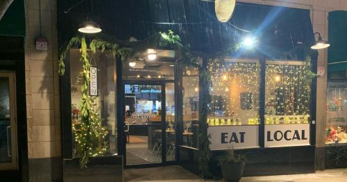 A cozy restaurant entrance with "EAT LOCAL" sign, warm lights, and greenery, inviting customers inside.