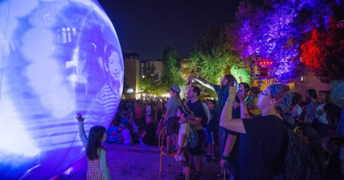 A crowd enjoys a nighttime outdoor event, watching projections on a large sphere surrounded by colorful lights.