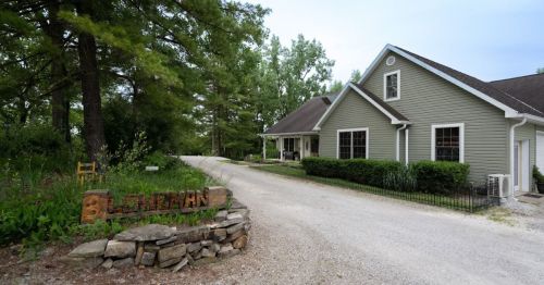 A gravel driveway leads to a green house surrounded by trees and shrubs, with a wooden sign reading "Graham."