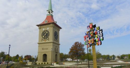 A clock tower with a green spire stands beside a colorful coat of arms display in a park-like setting.