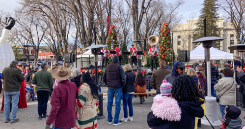 A festive outdoor event with a band performing on stage, surrounded by a crowd and decorated trees.