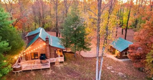Aerial view of a cozy log cabin surrounded by autumn trees, with a deck and string lights glowing at sunset.