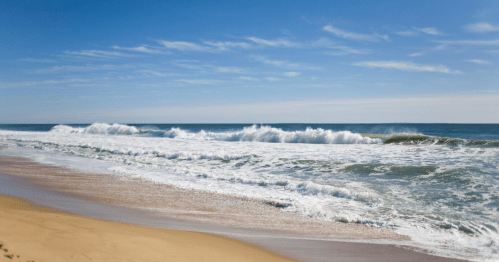 Waves crashing on a sandy beach under a clear blue sky with scattered clouds.