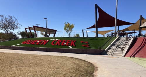 A park in Queen Creek, AZ, featuring green turf with the words "QUEEN CREEK AZ" and colorful shade structures.