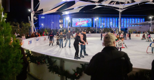 A lively ice skating rink with people skating, surrounded by festive decorations and a large screen in the background.