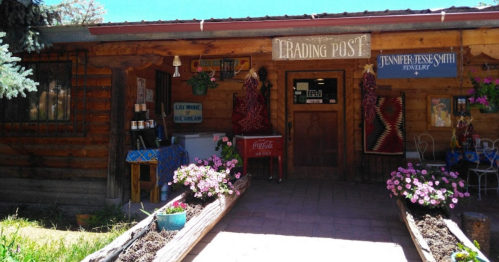 A rustic trading post with a wooden exterior, flowers, and signs for jewelry and ice cream.