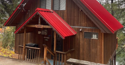 A wooden cabin with a red metal roof, surrounded by trees, featuring a porch and a grill.