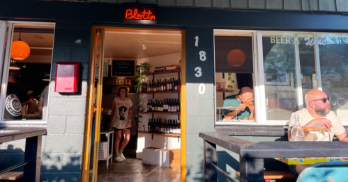 A cozy wine bar entrance with shelves of bottles, a server, and patrons enjoying food outside on a sunny day.