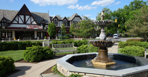 A decorative fountain in a park with benches, surrounded by greenery and Tudor-style buildings in the background.