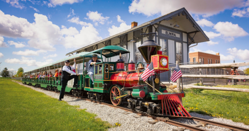 A colorful miniature train with passengers, parked near a historic train station under a blue sky with fluffy clouds.