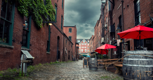A cobblestone alley lined with red umbrellas, brick buildings, and greenery under a cloudy sky.