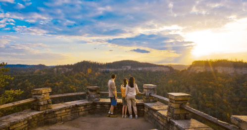 A couple stands on a stone overlook, gazing at a sunset over a lush, mountainous landscape.