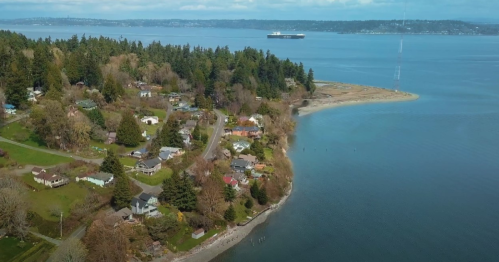 Aerial view of a coastal town with homes, trees, and a calm blue bay under a clear sky.