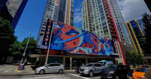 A vibrant cinema marquee with colorful artwork, surrounded by tall buildings and busy city streets.