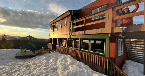A mountain cabin with a deck, surrounded by snow, and a tent nearby. A person stands on the deck enjoying the view.