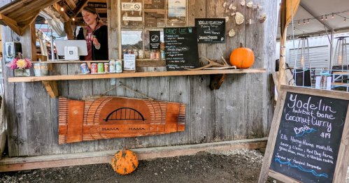 A rustic food stand with a menu board, a smiling vendor, and a carved wooden sign, featuring a pumpkin and drinks.