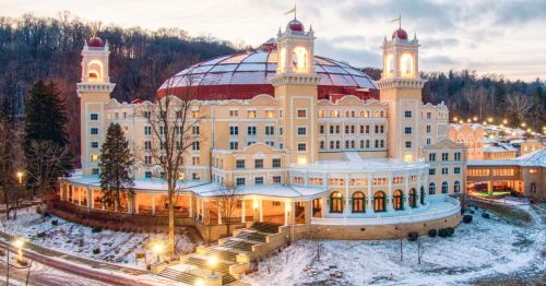 A grand hotel with a domed roof, surrounded by trees and snow, illuminated in the evening light.