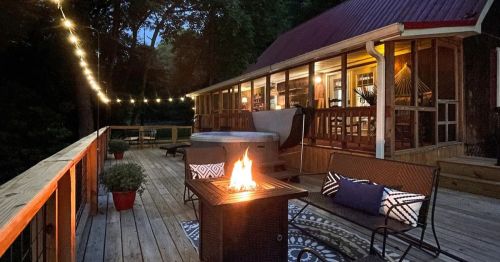 Cozy outdoor deck at dusk with a fire pit, string lights, and a hot tub, surrounded by greenery.