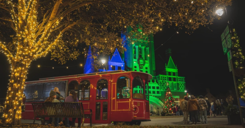 A festive scene at night with a red trolley, decorated trees, and a brightly lit building in green and blue.