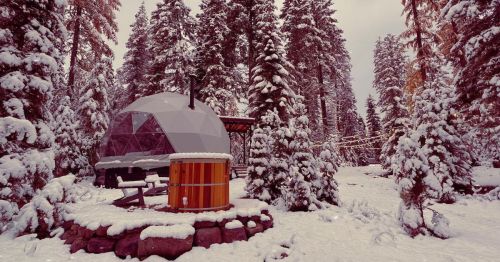 A geodesic dome cabin surrounded by snow-covered trees, with a wooden hot tub nearby in a winter landscape.