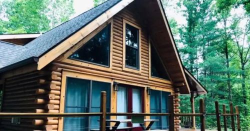 A cozy log cabin with a triangular roof, surrounded by lush green trees and a wooden deck.