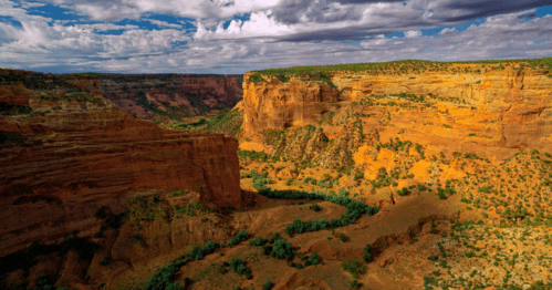 A stunning canyon landscape with red rock formations, green vegetation, and a winding river under a cloudy sky.