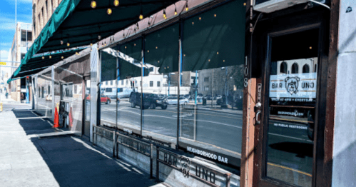 Exterior of a neighborhood bar with large windows, green awning, and a sign indicating public restrooms.