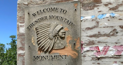 A weathered sign reading "Welcome to Thunder Mountain Monument" with a carved Native American chief's head.