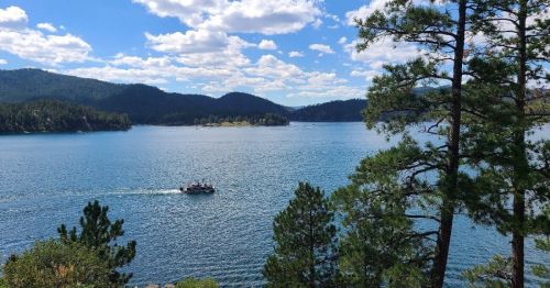 A serene lake surrounded by mountains and trees, with a boat gliding across the water under a blue sky with clouds.