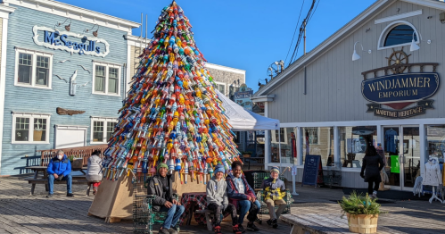 A colorful buoy tree stands in a coastal area, with people sitting nearby and shops in the background.