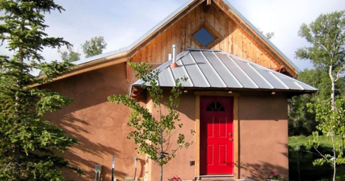 A small, rustic house with a red door, wooden accents, and a metal roof, surrounded by greenery.