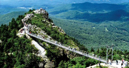 A scenic view of a rocky mountain with a suspension bridge and visitors, surrounded by lush green forests and hills.