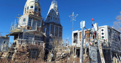 Whimsical, eclectic buildings with unique architecture, surrounded by a clear blue sky and overgrown vegetation.