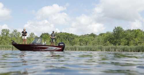 Two people stand on a fishing boat in a calm lake surrounded by lush greenery and blue skies.