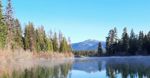 A serene lake reflects trees and mountains under a clear blue sky, with mist rising from the water's surface.