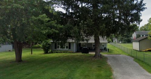 A single-story house with a large tree in the front yard, surrounded by green grass and a gravel driveway.