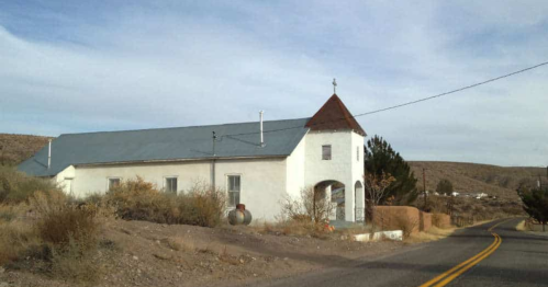 A white church with a gray roof sits beside a rural road, surrounded by sparse vegetation and hills in the background.