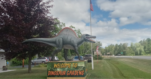 Sign for Dinosaur Gardens featuring a large Spinosaurus statue, with trees and a flag in the background.