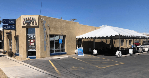 A tan building with a tent for outdoor dining, featuring a sign that says "Open for Take Out" and a blue sky above.