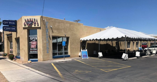 A tan building with a tent for outdoor dining, featuring a sign that says "Open for Take Out" and a blue sky above.