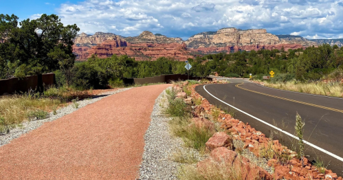 A scenic view of a winding road alongside a gravel path, with red rock formations and greenery in the background.