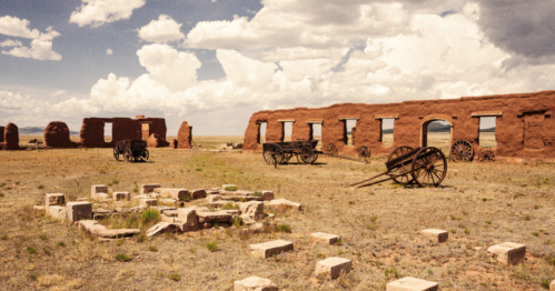 Ruins of a historic building with old wagons in a grassy landscape under a cloudy sky.