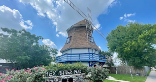 A windmill structure at Tex Ritter Park, surrounded by greenery and flowers under a blue sky with clouds.