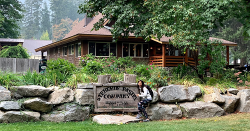 A person sits on a rock near a sign for Stehekin Pastry Company, with a rustic building and greenery in the background.