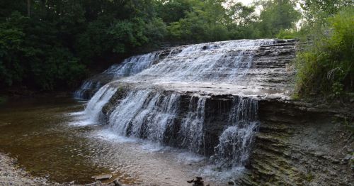 A serene waterfall cascading over rocky ledges into a calm stream, surrounded by lush greenery.